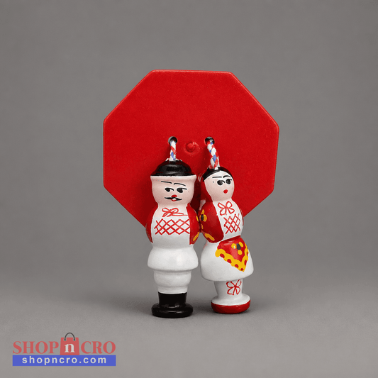 A pair of traditional Russian wooden Christmas ornaments shaped like a bride and groom, standing in front of a red octagonal backdrop. The groom figure wears a white robe with a red lattice pattern and black shoes, while the bride figure wears a red and yellow dress. Traditional Couple Under Šestine Umbrella Ornament