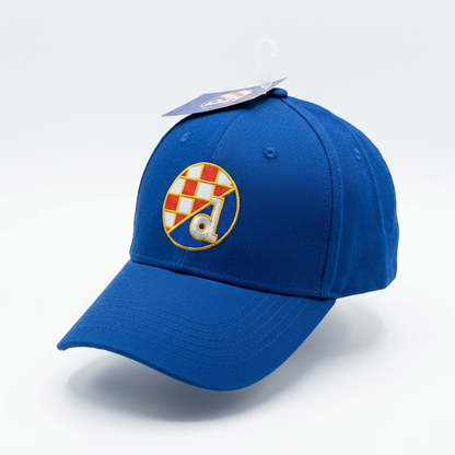 A royal blue baseball cap with a circular embroidered crest on the front featuring a red and white checkered pattern, a gold border, and a white letter 'a'. The cap is shown on a white background with a plastic hanger tag attached. Baseball Cap Dinamo Zagreb in Blue shopncro