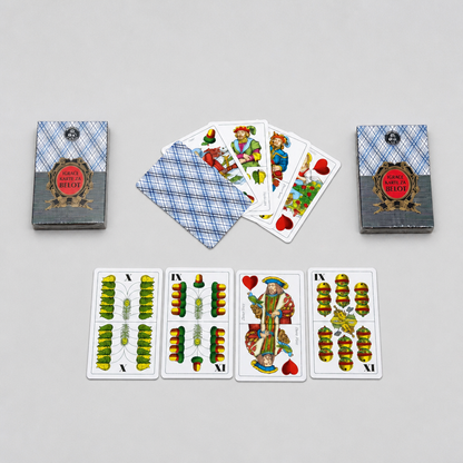 A set of playing cards and two matching boxes arranged on a light gray background. The cards show illustrated faces and backs with a blue plaid pattern, and the boxes have a gray and blue design with a red circular label. Croatia BELOT Playing Cards