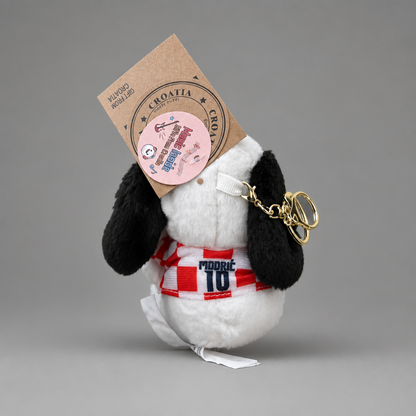 A small plush dog keychain photographed on a smooth gray studio background. The plush is mostly white with black ears and a red-and-white striped scarf featuring the text 'Modric 10'. Singing Croatian Fan Puppy – Hanging Plush Toy