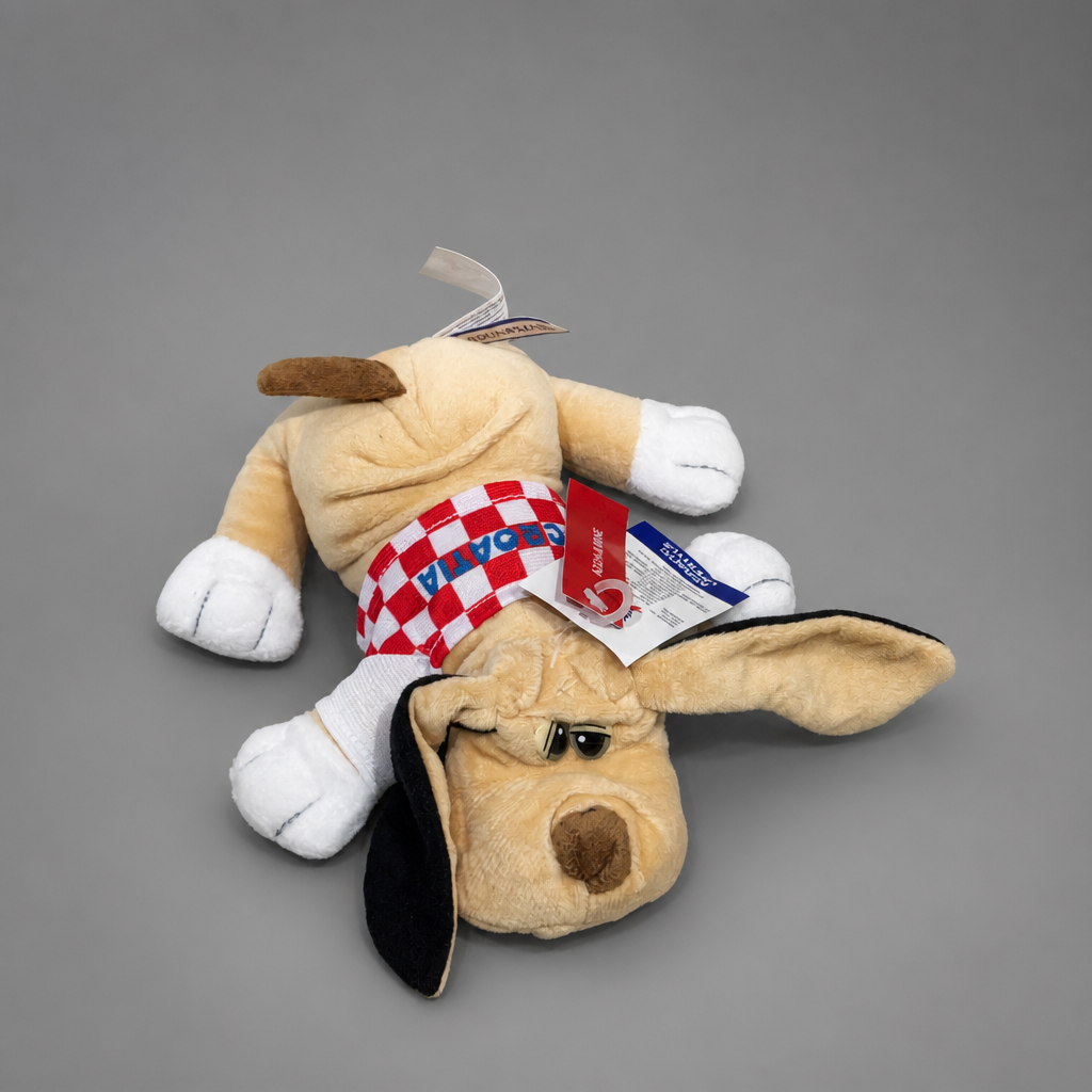 A plush dog toy lying on its side against a plain gray background. The toy is tan with white paws and feet, a black ear, brown nose and ear tip, and a red-and-white checkered scarf. Singing Croatian Fan Puppy – Plush Toy