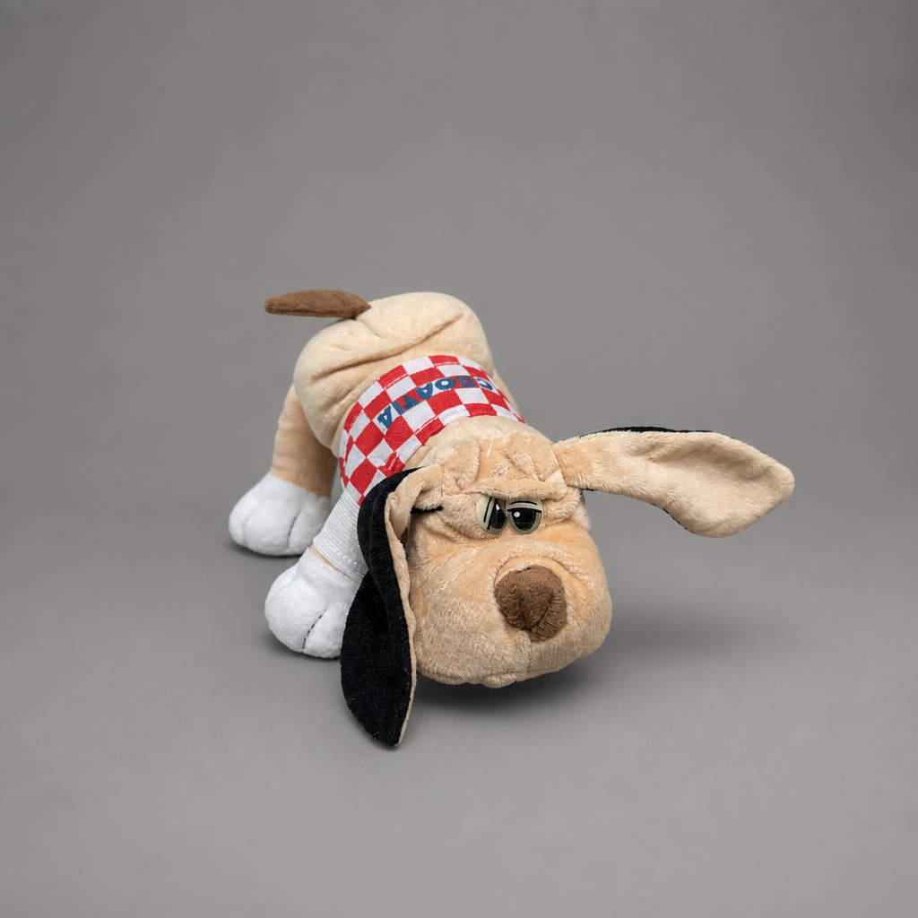 A plush dog toy lying on its side against a solid gray studio background. The toy is tan with long floppy black ears, white paws, a brown nose and tail, and wears a red-and-white checkered bandana with blue details. Singing Croatian Fan Puppy – Plush Toy