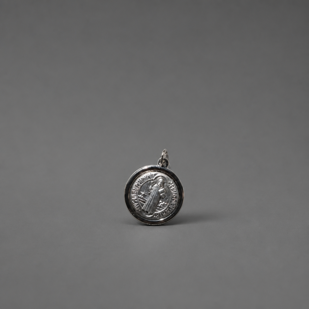 St. Benedict Medal | Pendant + Necklace or Pendant only A small round silver pendant or charm with an embossed figure and text, photographed on a smooth dark gray background.